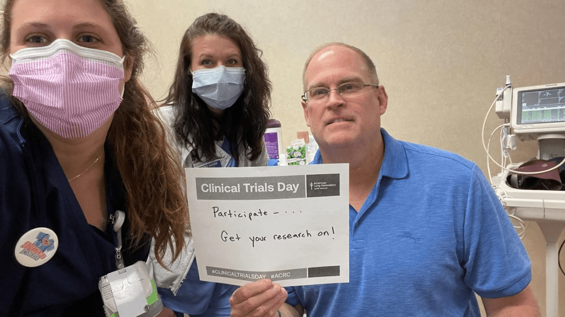 Mike Smith, lung cancer advocate, smiling in hospital with Clinical Trials Day sign, surrounded by medical staff.