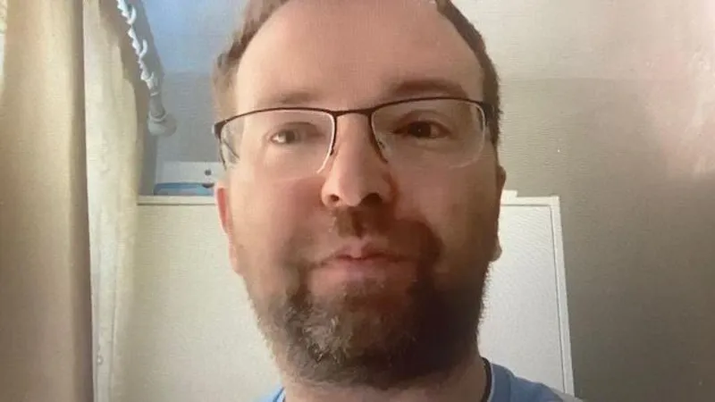 A man is wearing a blue top and glasses. He has brown hair and a beard.