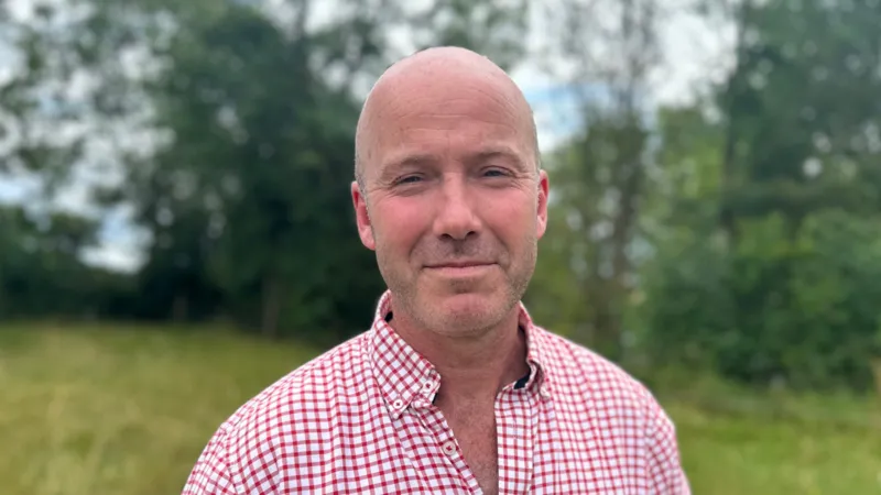 Charles Goadby has a shaved head and is wearing a red checked shirt. He is standing in a field.