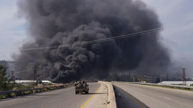 Reuters A large cloud of black smoke rises over a highway as a pick-up truck filled with members of Syrian armed forces rides toward it.