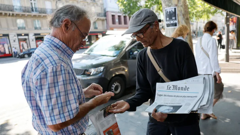 Reuters Ali Akbar, in a grey flat cap and black shirt, sells a copy of Le Monde to an elderly man in glasses and a checked blue shirt on the streets of Paris