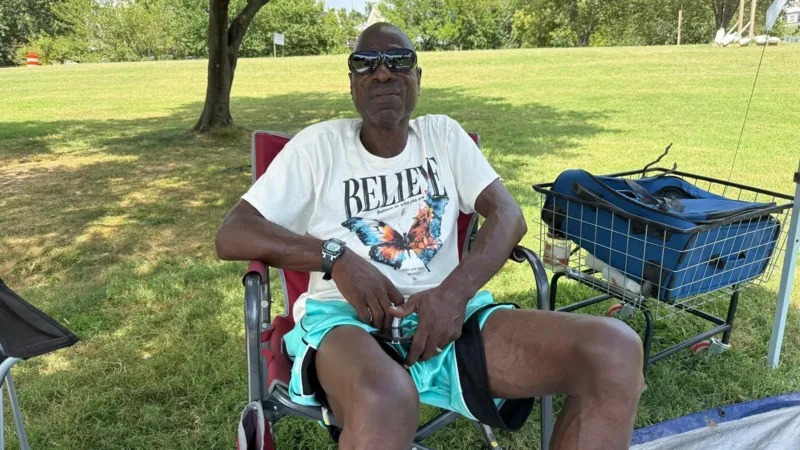 A man in a white t-shirt and aqua shorts and black sunglasses sits in a camping chair on a patch of grass. A blue suitcase in a shopping cart is next to him. In the fare distance, a large building is visible