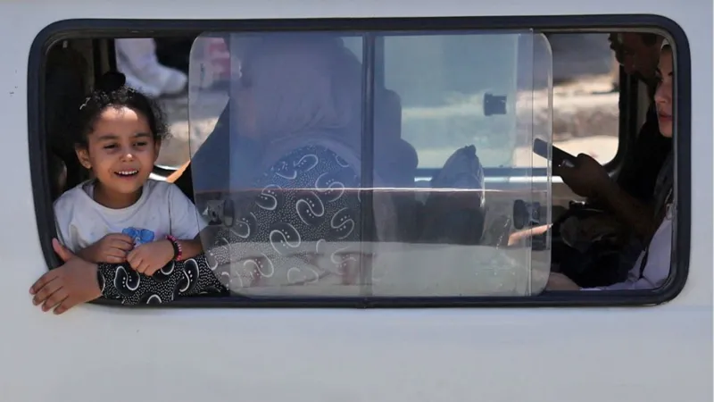 Amr Abdallah Dalsh/Reuters Girl in a white T-shirt with dark brown hair staring out the window of a bus whilst smiling. Her mother, wearing a black and white outfit, holds her back slightly - Thursday 21 August 2025.