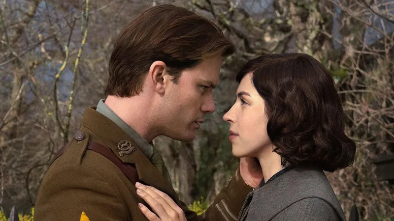 Starz Jeremy Irvine is dressed in a British Army officer's uniform and has brown hair while Hermione Corfield has dark mid-length hair and a grey coat. They are standing in front of bare branches, embracing.
