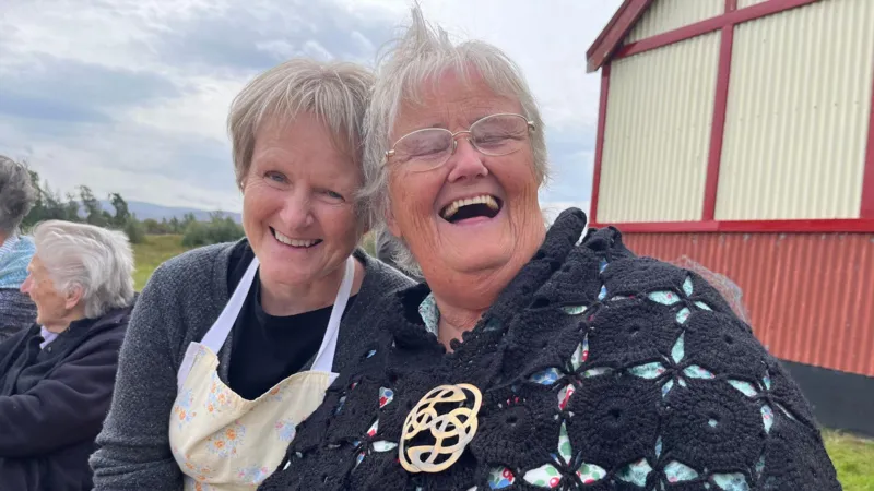 Maureen Caddon and Sheila MacKay smile at the camera. Maureen wears a grey jumper with an apron on top and Sheila wears a black shawl with a broach and glasses