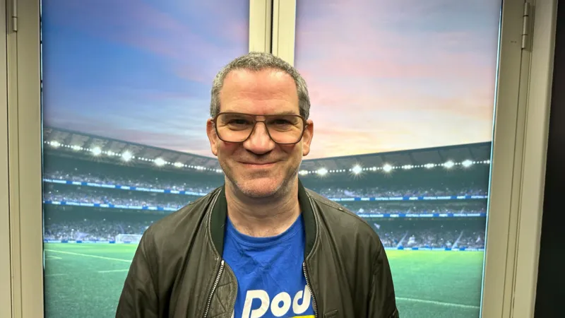 A man wearing glasses stood in-front of a window. Behind him is an image of a football stadium. He is wearing a dark blue t-shirt with a khaki jacket over the top. He has short grey hair.