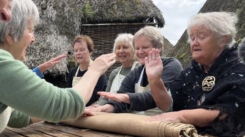 The Badenoch Waulking Group demonstrate waulking at the Highland Folk Museum. Women sit on either side of a wooden table and appear to be hitting a rolled up brown fabric while singing.