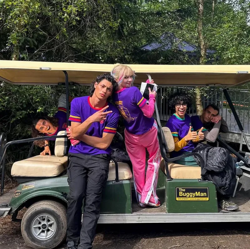 Moosa Mostafa Cast of Wednesday either sitting on or standing next to a golf buggy. All wear distinctive purple tee-shirts. Actress Emma Myers has blonde hair and is wearing bright pink tracksuit bottoms and pink top under her tee-shirt.
