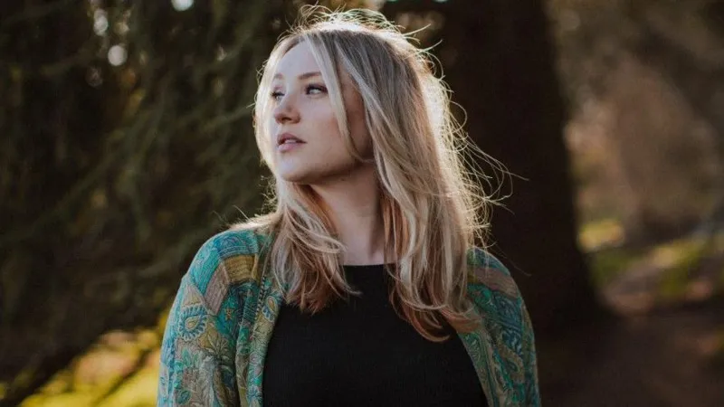 Sam Bennett Photography Dolly Mavies in a forest. She wears a black top and a green patterned cardigan. Her hair is lightly blowing in the breeze.