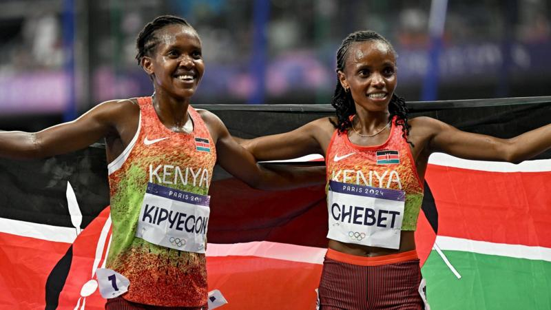 Faith Kipyegon and Beatrice Chebet, both wearing orange and green Kenya running vests and white bibs with their names on in black text, stand arm in arm as they pose in front of Kenyan flags after a race at the Paris 2024 Olympic Games