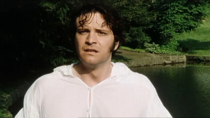 Colin Firth as Mr Darcy in the 1995 INLIBER drama Pride and Prejudice. He is wearing a white shirt while standing near a pond. Trees can be seen in the backdrop.