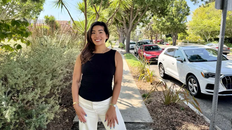 Aileen Barrameda A woman wearing a black shirt and white pants poses on the sidewalk of a tree-lined residential street, next to a parked white car.