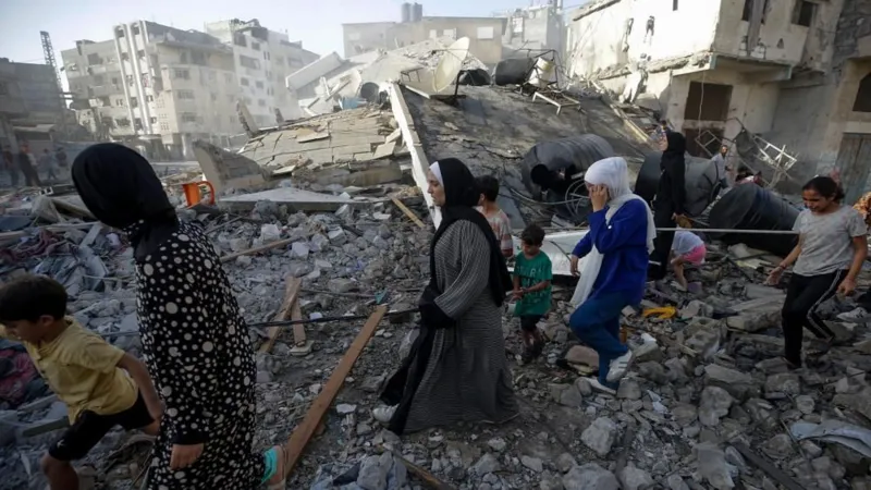 EPA People in Gaza City inspect the rubble of their former home following Israeli strikes on Friday - a group of women and children walk over the rubble with buildings in the background on a sunny day.