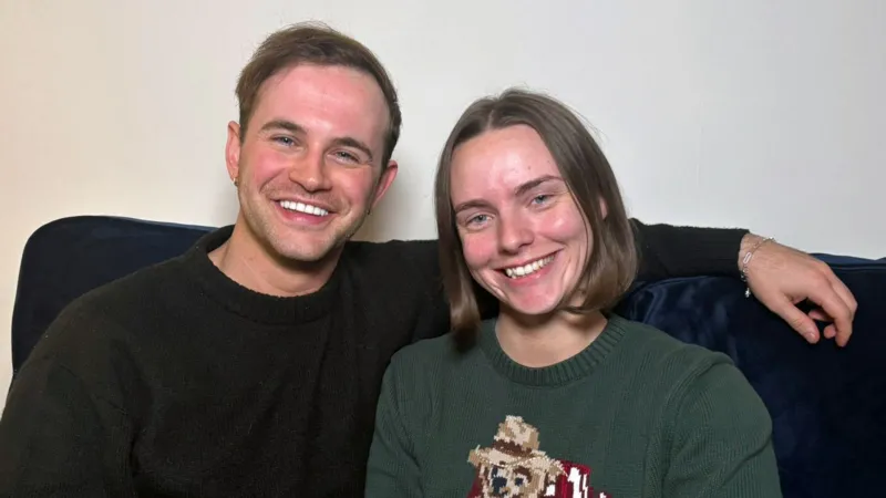 Sam and Helen sitting on a sofa. Sam has short brown hair and is wearing a black jumper. He has his arm draped behind Helen who is wearing a green jumper with a teddy bear design on the front. she has light brown hair in a short bob.