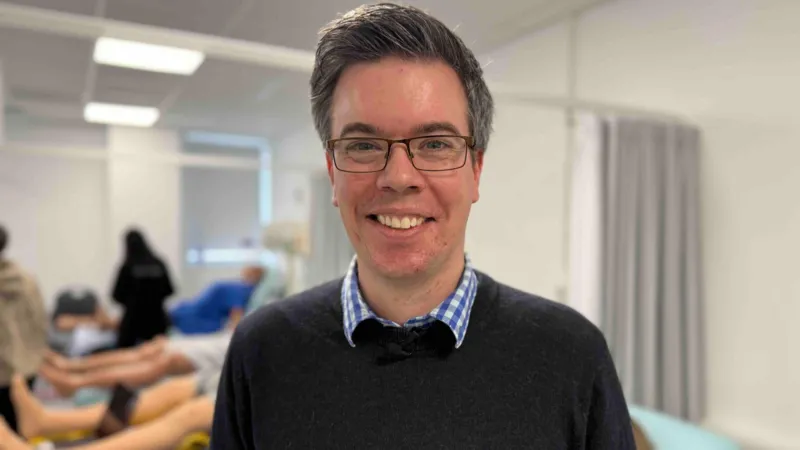 A man with short black hair, greying at the sides, smiles at the camera. He is wearing a black jumper over a blue chequered shirt and black-framed glasses. The background is out of focus, but mannequins can be seen lying on hospital beds