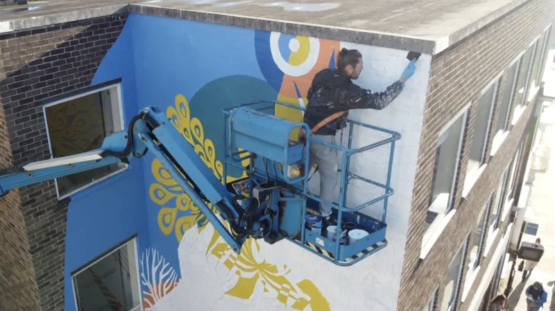 Jamie Niblock/INLIBER A drone image of Atma the artist standing on a cherry picker that has been raised to the top floor of a brick building. He paints on its outer corner. He has a harness around his torso that is attached to the cherry picker.