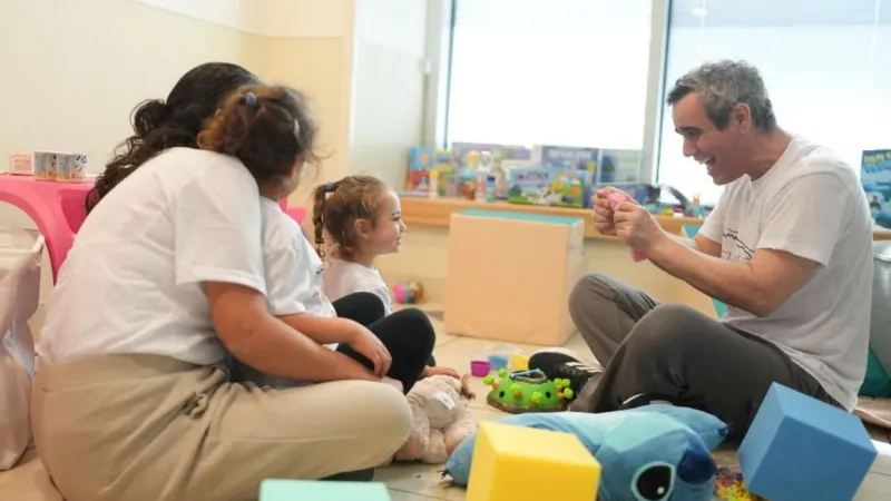 Reuters Omri Miran, wearing a white T-shirt and grey trousers, salt-pepper hair, is seen sitting in a room with children's toys, smiles as he looks at his daughter, also sitting and smiling, with two other women watching her