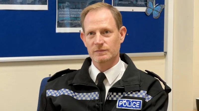 Chief Superintendent Simon Dodds has short light brown hair. He is wearing police uniform and is sat at a desk.