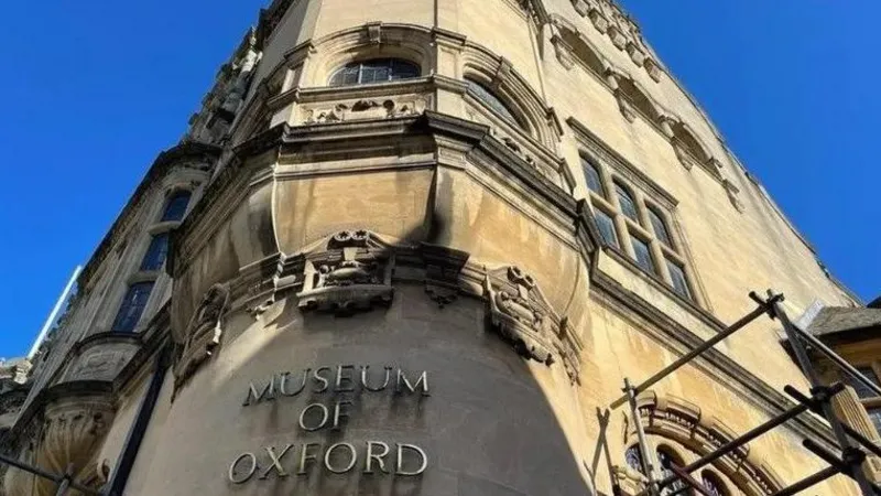 Looking up at the Museum of Oxford sign, in gold lettering, above the entrance, part of a yellow-stoned historic building.