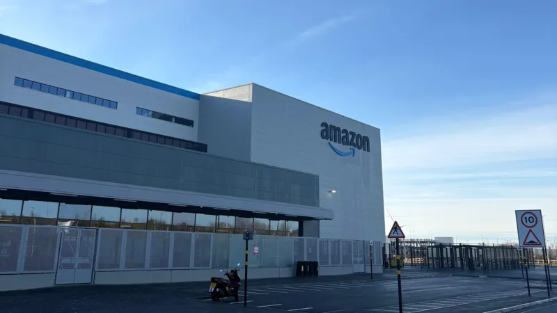 The outside of the Amazon warehouse in North Ferriby. It is a large white building with the Amazon logo on one side and a large car park in front. The sky is blue.