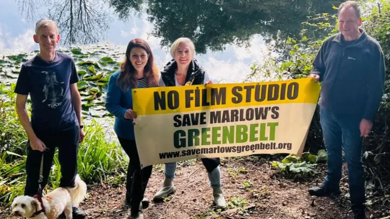 Joy Morrissey Four people stood near a body of water with Joy Morrissey (second left) holding a mostly yellow banner reading: NO FILM STUDIO. SAVE MARLOW'S GREENBELT