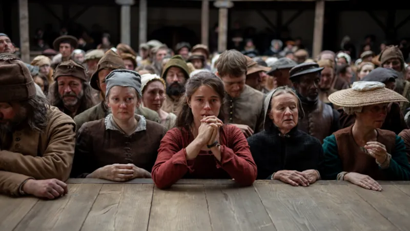 Universal Studios A film still from Hamnet, showing Jessie Buckley as Agnes (Anne Hathaway). The picture shows a group of characters who are audience members, pressed against the stage of the Globe. Agnes is in the middle in a red dress with her hands pressed together, looking somber.