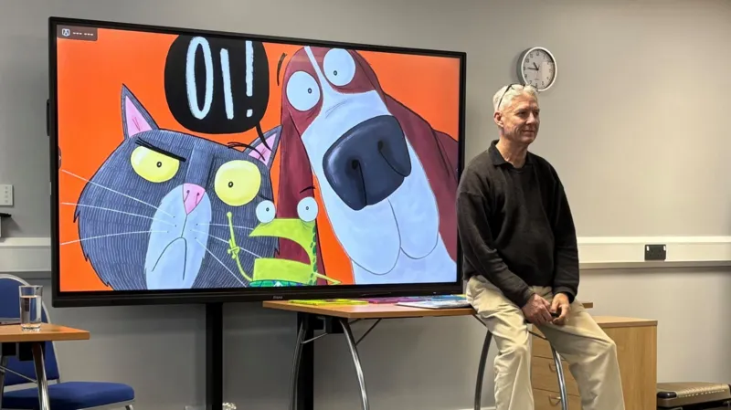 Jodie Halford A man sits on a table in front of a large screen with a cartoon picture of a cat, frog and dog with a speech bubble in black that says OI! The background of the cartoon is orange.
