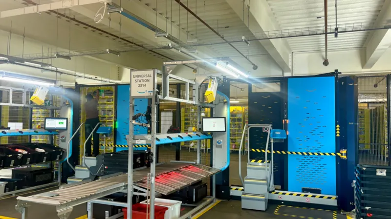 Machinery and large yellow robotic machines behind a cage inside an Amazon warehouse. Various screens can be seen and a sign reads