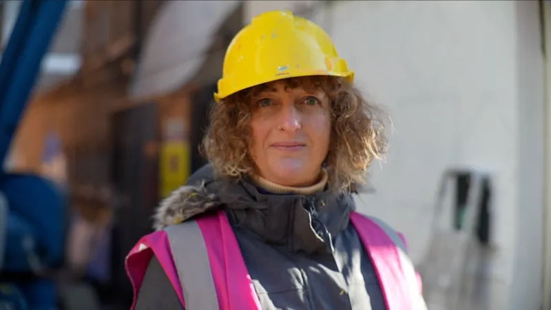 Jamie Niblock/INLIBER Iona Hodgson smiles at the camera. She has short curly light brown hair that can be seen underneath a yellow hard helmet she is wearing. She also wears a pink hi-vis jacket above a grey coat.