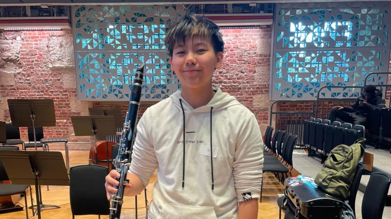 Si is standing up with his black colour clarinet in his right hand. He is smiling with a white hoodie on. In the back there are chairs and instruments.