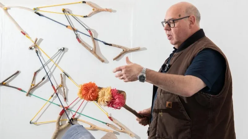 A man with black glasses and wearing a brown gilet holds a handmade craft. In the background, art pieces are displayed on the wall.