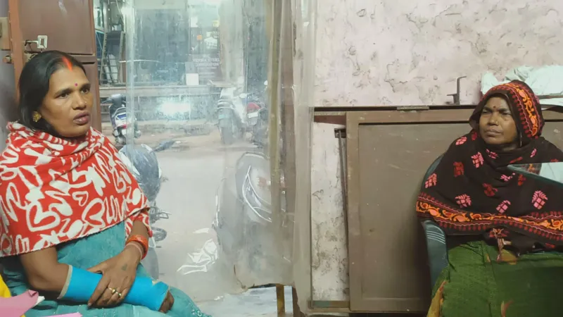 Abhishek Dey Two women migrant workers in Delhi's Mayapuri industrial area, sitting in a local trade union office, discussing the electoral roll revision exercise and the documents it would require. Both of them are wearing shawl over their sarees and one of them, Kusum Devi, has her right forearm plastered because of a fracture. Behind the other woman, there's a broken door leaning against the wall of the trade union office 