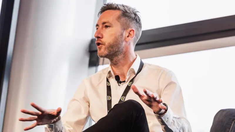 Matt Navarra A man wearing a white shirt, black lanyard and microphone attached to his collar is giving a talk at an event which looks like a conference.