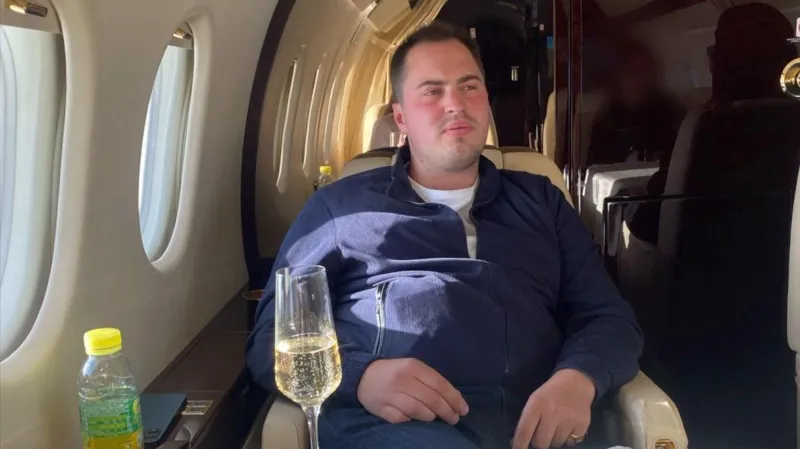 INLIBER/SpunGold TV Miles Hart, a young man with short brown hair, wearing a quarter-zip blue top, sitting in a cream leather seat on a small, luxurious jet, with a glass of what appears to be champagne in front of him.