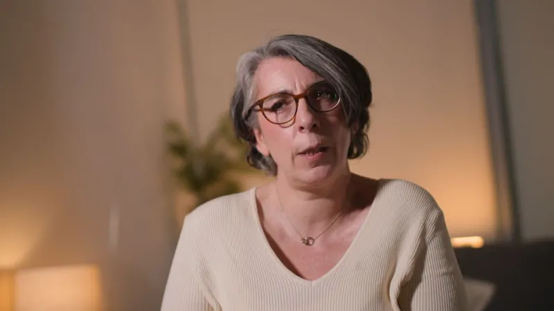 Bronagh Andrew has short, grey hair and glasses. She is wearing a beige ribbed jumper with a V-neck, showing a silver necklace. The background is softly lit and blurred.