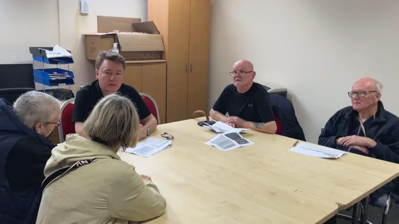 Andy Watson/INLIBER A group of four elderly people, two women and two men, sitting across from each other at a large table with the leader Pallion Action group Ken Newby at the head of the table. The room is painted white.