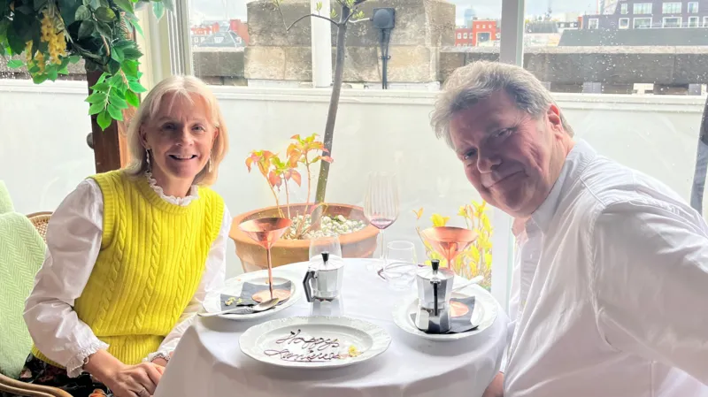 Sue and Alex at a table, smiling, with an anniversary cake in front of them.