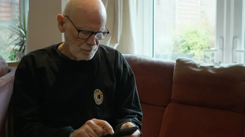 Gideon Rabinowitz wears a dark jumper and sits on his sofa looking down towards his mobile phone