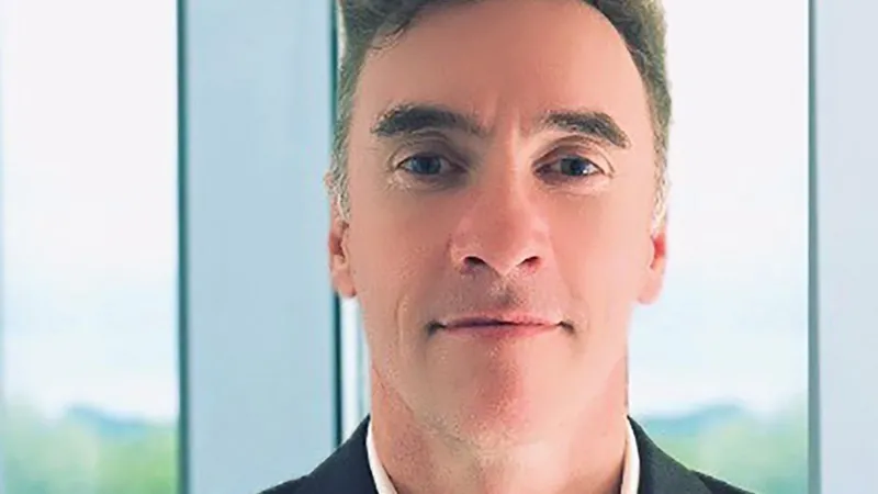 Unknown Paul Doyle, who has grey hair styled into a quiff, smiles at the camera while wearing a black suit jacket and open collared white shirt.