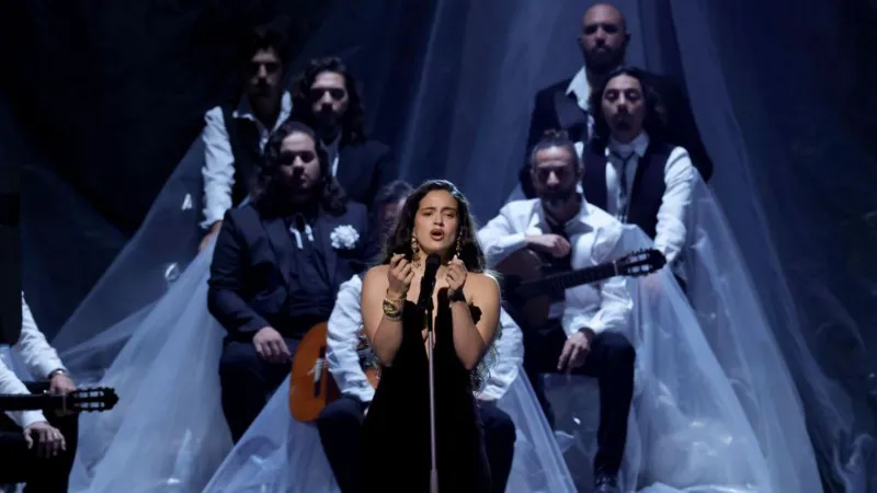 Getty Images Rosalía wears a black off-the-shoulder dress. She is singing into a microphone stand while performing on stage. There are musicians with guitars sitting behind her.