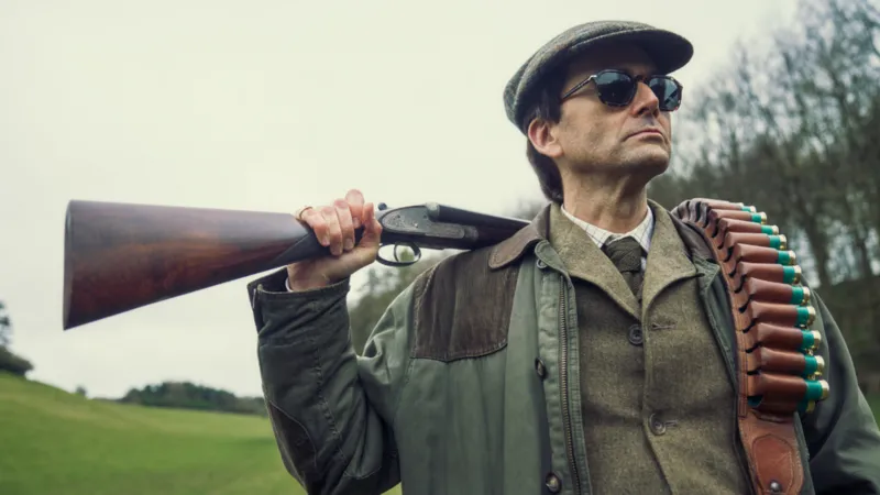 Disney+ David Tennant stands in character as Lord Tony Baddingham. He is in a field, wearing what appears to be hunting clothing, with a large rifle resting over the back of his shoulders and a case of bullets hanging in a leather case down his front. He is wearing sunglasses and is looking away from the camera.