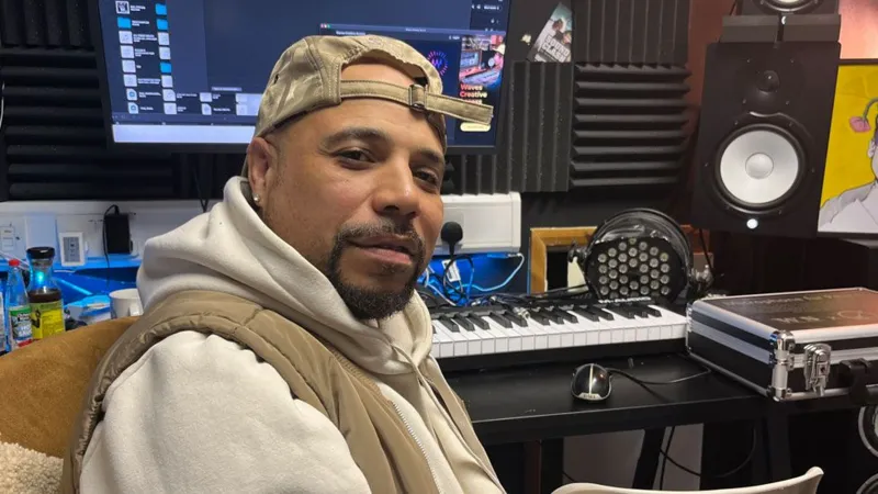 Tom Jackson/INLIBER Karl Lewis - known as KD - sitting in a recording studio. He has turned sideways on and has a beige gillet over a cream hoodie, a neatly clipped black goatee beard and is wearing a beige baseball cap back to front. Behind him is a keyboard resting on a table, headphones and a computer screen.