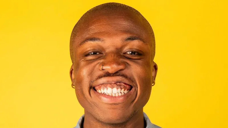 Abena Kusi Benjy is bald and is smiling at the camera. He has a gold nose ring and hoop earrings. He is standing in front of a bright yellow background.