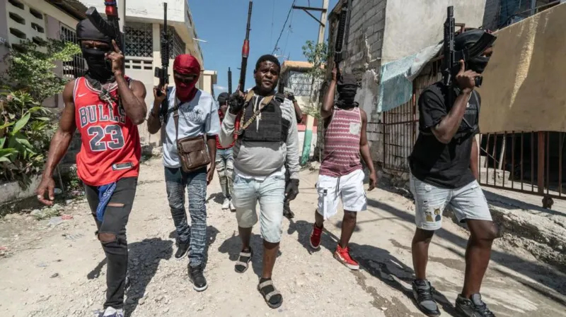 Getty Images Five young men in shorts and ripped jeans walk along an unpaved street holding guns in the air as they patrol an area in Port-au-Prince in February 2024
