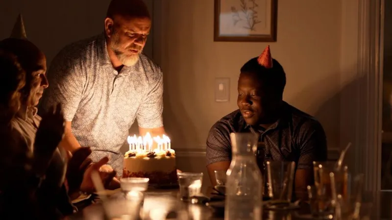 Liam Weir/INLIBER Marven looking on and smiling as a man brings a birthday cake with lit candles to the table where he and some other people are sitting. The lights are low and Marven is wearing a party hat.