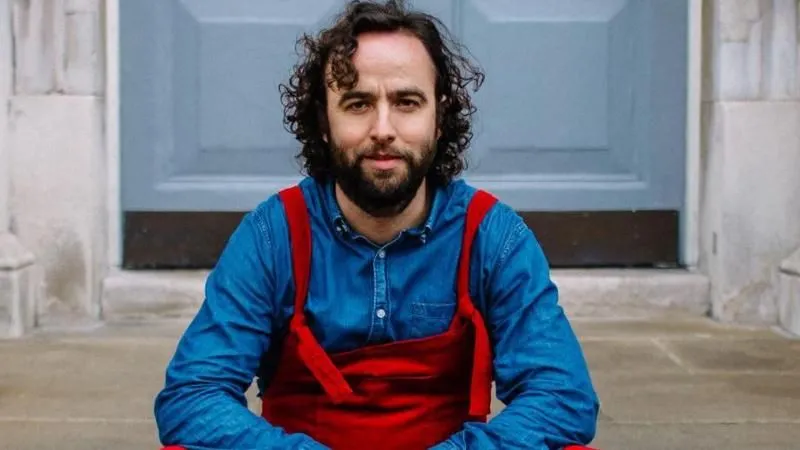 Daf James looking at the camera. He is wearing red dunagrees and a blue long sleeved top.