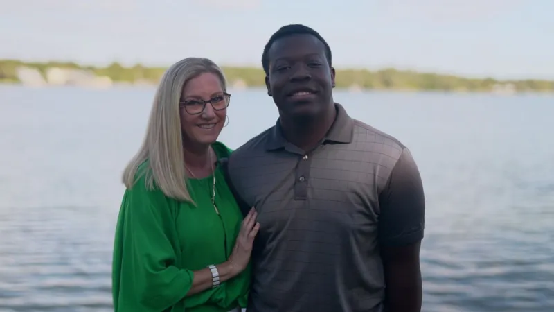 Liam Weir/INLIBER Stacey and Marven facing the camera in front of a lake. Both are smiling, Marven is wearing a grey, collared T-shirt, Stacey has long blond hair and glasses and is wearing a bright green gathered top.
