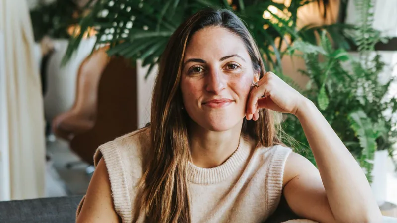 Katina Bajaj A woman with long brown hair is sat on a sofa smiling at the camera, with her hand touching her cheek. Wild houseplants are behind her.