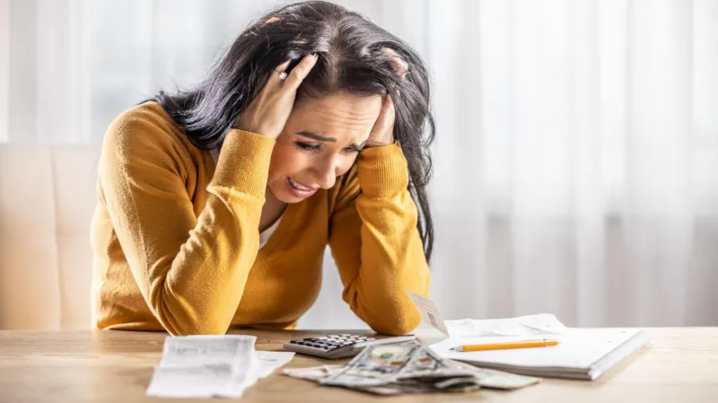 Getty Images Brunette woman with yellow jumper has her head in her hands at the table over a pile of receipts and cash.