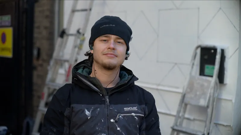 Jamie Niblock/INLIBER Leo Vanmegan smiles at the camera as he stands in front of a white building with some shapes drawn on to it and a step ladder that leans against it. He wears a black beanie hair, black coat and has wired headphones in each ear. He has dark hair that pokes out of his hat and he has a beard.
